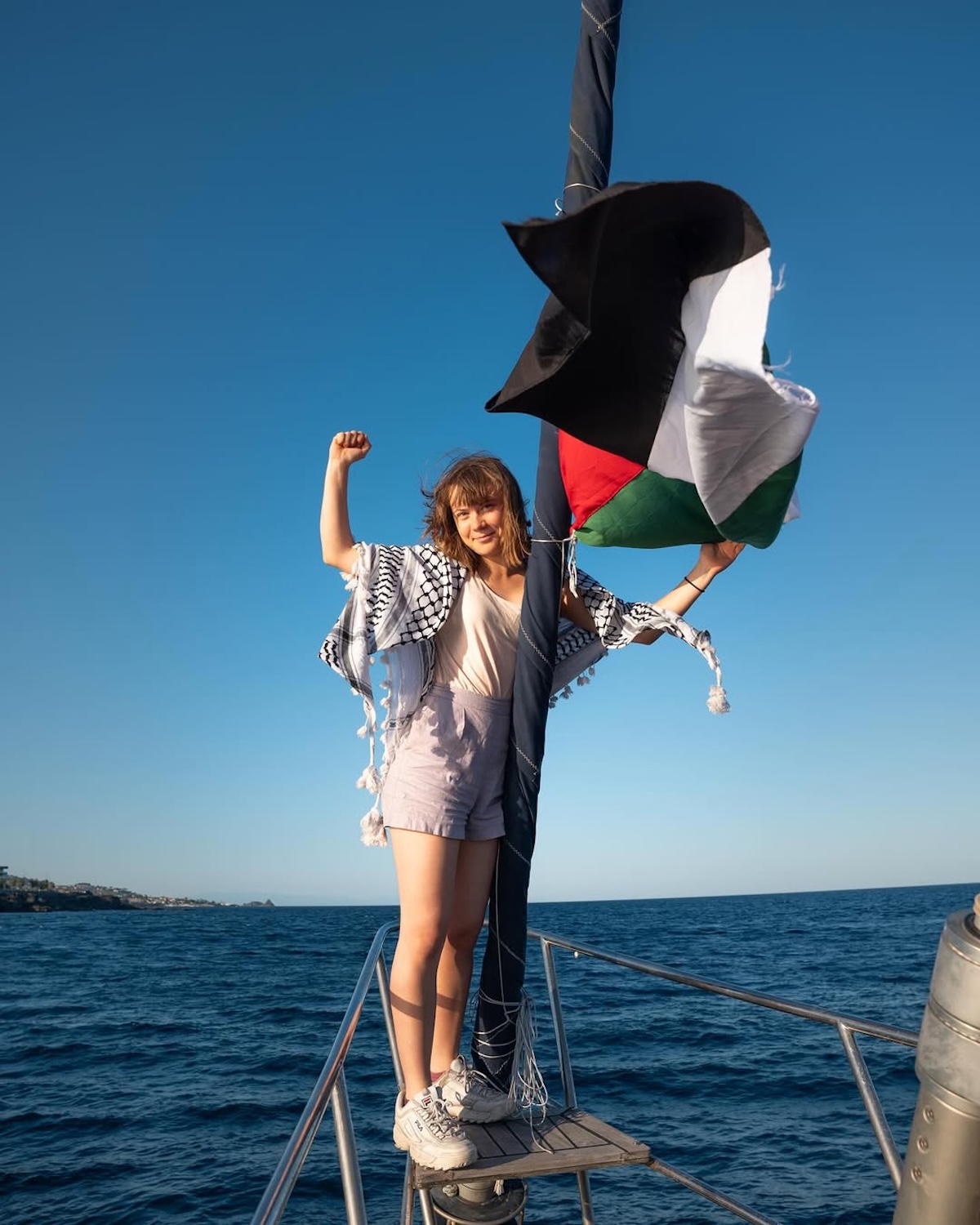 Greta Thunberg Gaza: Dziewczyna w jasnych szortach, białym topie i z chustą palestyńską przewieszoną przez ramiona stoi na dziobie jachtu, trzymając się masztu, do którego przywiązana jest flaga Palestyny. Unosi zaciśniętą pięść w geście protestu. Tło stanowi spokojne morze i jasne niebo.