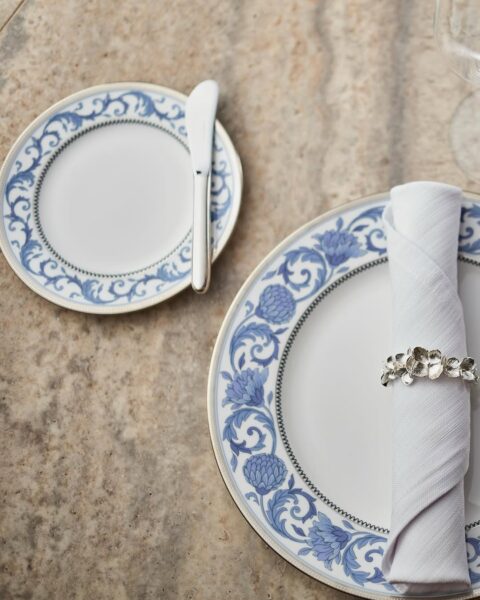 Zbliżenie na eleganckie nakrycie stołu. Na marmurowym blacie leży porcelanowy talerz ozdobiony niebieskim wzorem inspirowanym chińską ceramiką. Na talerzu znajduje się zwinięta biała serwetka, przewiązana srebrnym pierścieniem z motywem kwiatów. Obok mały talerzyk z tym samym wzorem, na którym leży smarownica do masła. Całość tworzy harmonijną, klasyczną i wysmakowaną kompozycję.