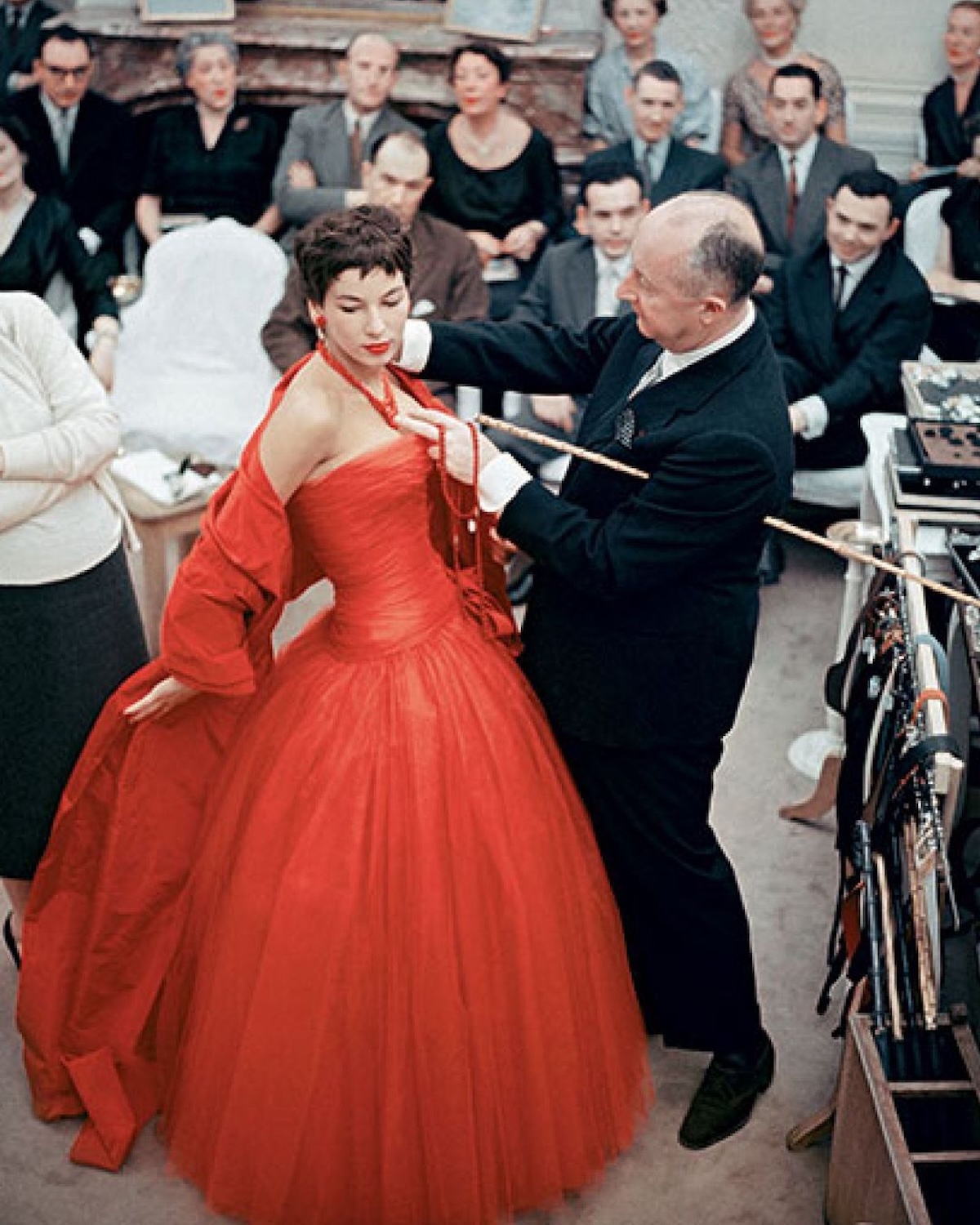 Dior wystawa 2025: Kolorowa fotografia historyczna ukazująca Christiana Diora w trakcie przymiarki czerwonej sukni balowej na modelce. Scena rozgrywa się w atelier w obecności zespołu i dziennikarzy, dokumentując proces twórczy oraz intensywną pracę nad detalami – charakterystyczną dla Diora precyzję i perfekcjonizm.