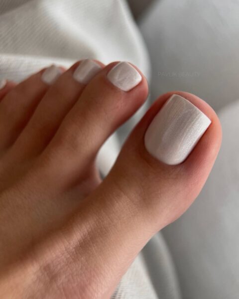 Zdjęcie stopy ze zbliżeniem na paznokcie. Widać tu perłowy, biały manicure na sierpień 2025. Stopa pozuje na tle białego materiału.
