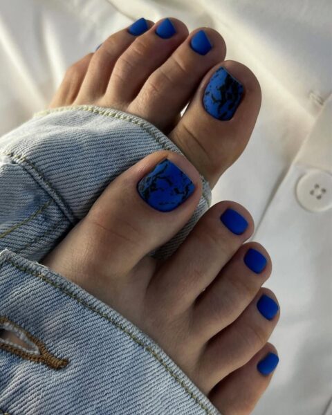 Zdjęcie stóp z granatowym pedicure w odcieniu borówki. Na dużych palcach użyto lakieru cracked nails z efektem popękanego lakieru. Stopy pozują w jeansowych nogawkach.