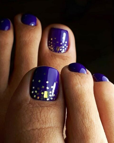 Zdjęcie stóp z granatowym pedicure. Duże palce są ozdobione dużymi płatkami brokatu w kolorze złotym. Stopy pozują na czarnym tle i prezentują pedicure.