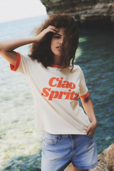 Modelka ubrana w kremowy T-shirt z kontrastującym, pomarańczowym napisem „Ciao Spritz” oraz oblamówkami przy rękawach i dekolcie, pozuje nad turkusowym morzem na tle wapiennego klifu. Ma lekko rozwiane włosy i nonszalancko przytrzymuje je dłonią, co podkreśla klimat włoskich wakacji z lekką nutą vintage. Stylizacja zestawiona z jasnymi jeansowymi szortami oddaje ducha kolekcji – beztroskiego, słonecznego i autentycznego.