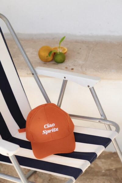 Pomarańczowa czapka z daszkiem z haftowanym napisem „Ciao Spritz” leży na pasiastym leżaku w granatowo-białe pasy, ustawionym na jasnym kamiennym tarasie. W tle cytrusy – pomarańcza, limonka i plasterek cytryny – przywołują klimat włoskiego lata i aperitivo. Stylizacja zdjęcia łączy wakacyjny luz z graficzną prostotą kolekcji She is Sunday.