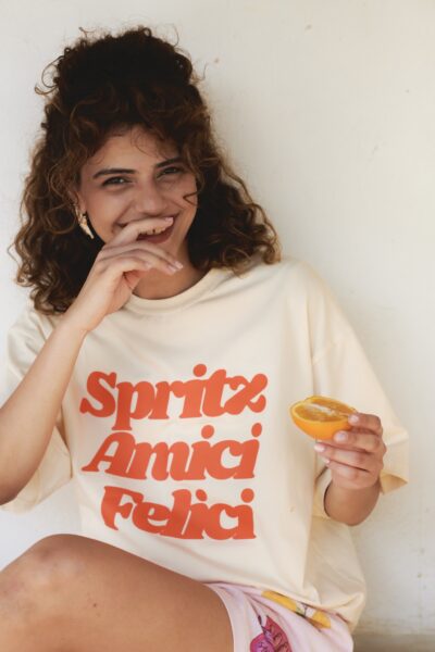 Uśmiechnięta kobieta w kremowym T-shircie z dużym, pomarańczowym napisem „Spritz Amici Felici” siedzi oparta o ścianę i trzyma w dłoni połówkę świeżej pomarańczy. Jej stylizacja uzupełniona jest pastelowymi szortami z owocowym printem, co nadaje całości figlarnego i wakacyjnego tonu. Naturalna fryzura i ekspresja twarzy oddają autentyczną radość i luz, wpisując się w estetykę codziennych przyjemności, które kolekcja celebruje.