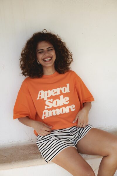Uśmiechnięta kobieta siedzi na kamiennej ławce, ubrana w luźny, pomarańczowy T-shirt z dużym napisem „Aperol Sole Amore” i krótkie szorty w biało-czarne paski. Stylizacja łączy letnią swobodę z mocnym akcentem graficznym. Zdjęcie uchwyca moment beztroskiej radości i luzu, będący esencją kolekcji „Spritz Season” od She is Sunday.