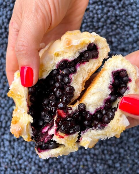 Zbliżenie na ręce z czerwonym manicure, rozrywające jagodziankę z lukrem, wypełnioną dużą ilością soczystych, całych jagód. W tle rozsypane świeże jagody tworzą kontrastowe tło.