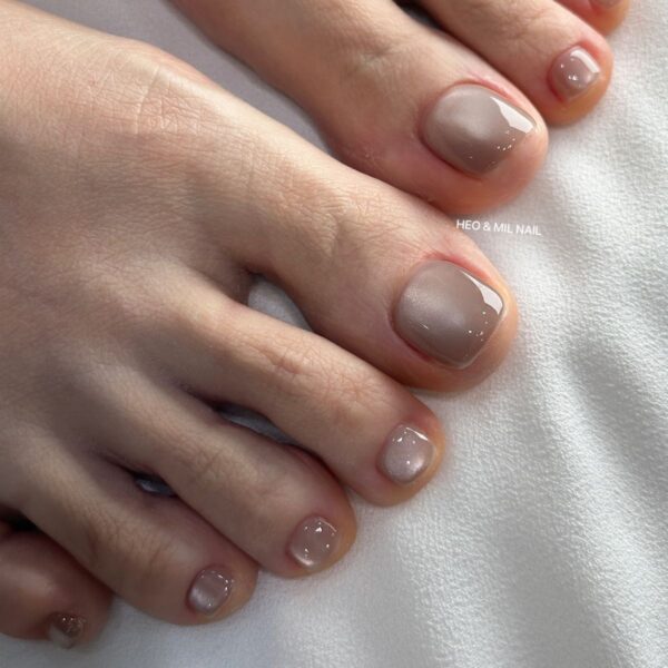 Subtelny pedicure w odcieniach chłodnego beżu i szarości, dający delikatny, elegancki efekt cieniowania. Paznokcie są krótkie i zadbane, pokryte błyszczącym topem. Tło stanowi biała tkanina.