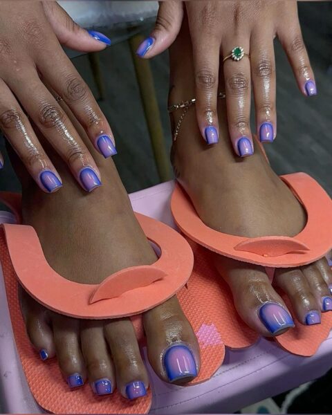 Zestaw manicure i pedicure w kolorze fioletowo-niebieskim z efektem ombre i wyraźnym połyskiem. Paznokcie są idealnie dopasowane do siebie. Stopy znajdują się w różowych japonkach do pedicure, a dłonie ozdobione są biżuterią – pierścionkiem z zielonym kamieniem.