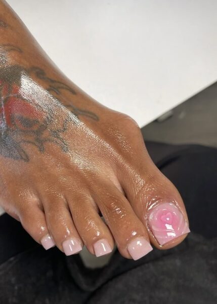 Pedicure w stylu minimalistycznym – jasnoróżowe, mleczne paznokcie z ozdobnym wzorem róży na dużym palcu. Stopa z widocznym tatuażem przedstawiającym kolorowy wzór, całość pokryta jest nawilżającym olejkiem, co nadaje połysk.