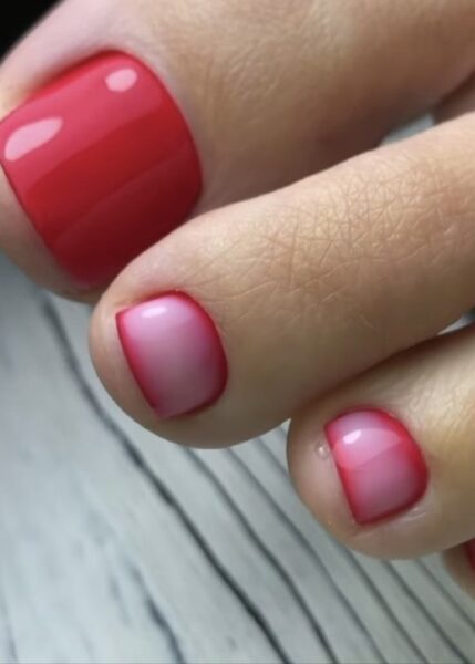 Intensywny pedicure z czerwonym dużym paznokciem i mniejszymi paznokciami w różowo-czerwonym ombre. Stylizacja ma bardzo nowoczesny i estetyczny wygląd, z wyraźną granicą między kolorami. Tło to jasne, drewnopodobne podłoże.
