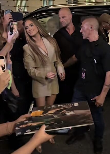 Zbliżenie na Jennifer Lopez podpisującą płytę dla fanów. Artystka ma na sobie elegancki beżowy komplet przypominający marynarkową sukienkę. Na twarzy widoczny pełny makijaż sceniczny oraz długie, proste włosy w stylu charakterystycznym dla Lopez.