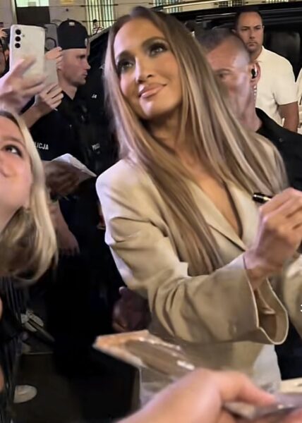 Gwiazda ubrana w beżowy garniturowy komplet o kroju mini, pozująca z uśmiechem do zdjęć w tłumie fanów przed wyjściem z samochodu. Otoczona przez ochroniarzy, rozdaje autografy i przyciąga uwagę klasyczną, a zarazem nowoczesną stylizacją wieczorową.