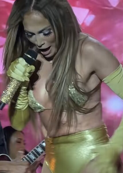Jennifer Lopez na scenie w połyskującym złotym kostiumie z rękawiczkami i mikrofonem obsadzonym kryształkami. Stylizacja sceniczna eksponuje sylwetkę i podkreśla estradowy charakter występu podczas koncertu w Warszawie.