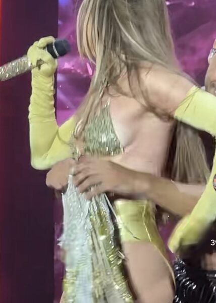 Jennifer Lopez na scenie w trakcie poprawiania elementów złotej stylizacji – frędzle i połyskujący materiał tworzą dynamiczny efekt sceniczny. Ujęcie pokazuje moment, w którym tancerz pomaga jej skorygować garderobę.