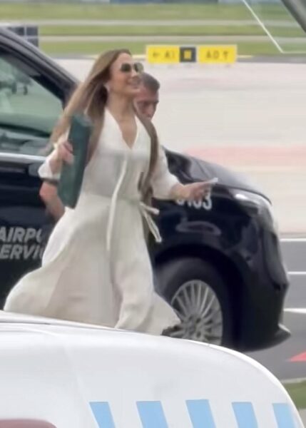 Jennifer Lopez uśmiechnięta i pewnym krokiem zmierzająca w kierunku wejścia do prywatnego samolotu. Ubrana w jasną, przewiewną sukienkę maxi oraz szpilki, prezentuje styl typowy dla modowego nurtu „airport chic”.