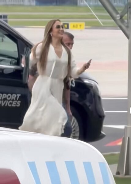Jennifer Lopez w lekkiej, zwiewnej sukience w kolorze kości słoniowej z wiązaniem w talii, zmierzająca do samolotu na płycie warszawskiego lotniska. W ręku trzyma zieloną kopertówkę, a na twarzy ma duże okulary przeciwsłoneczne.