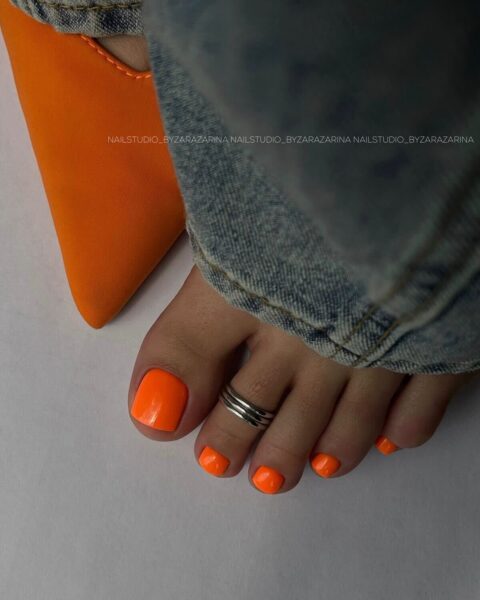 Neonowy pedicure w intensywnym, elektryzującym odcieniu pomarańczowym na tle dżinsów i dopasowanych butów w tym samym kolorze. Modna stylizacja paznokci u stóp wpisująca się w trend „Italian Summer”, idealna na lato 2025.