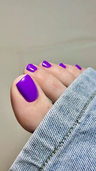 Kolory pedicure na lato 2025: Pedicure w intensywnym, głębokim odcieniu fioletu, z błyszczącym wykończeniem. Stylizacja nowoczesna i wyrazista, idealna na wieczorne wyjścia latem 2025. Paznokcie kontrastują z jasnym jeansem i naturalną karnacją.