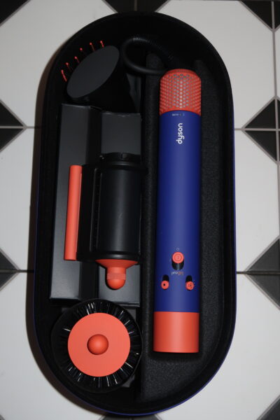 Multistyler Dyson Airwrap i.d.™ Straight+Wavy: Zestaw Dyson Airwrap i.d.™ Straight+Wavy w etui – widok z góry. W środku eleganckie akcesoria do stylizacji włosów w kolorach chabrowym i miedzianym: korpus urządzenia, końcówki, szczotki oraz elementy do suszenia i modelowania. Etui wyłożone czarną pianką ochronną.