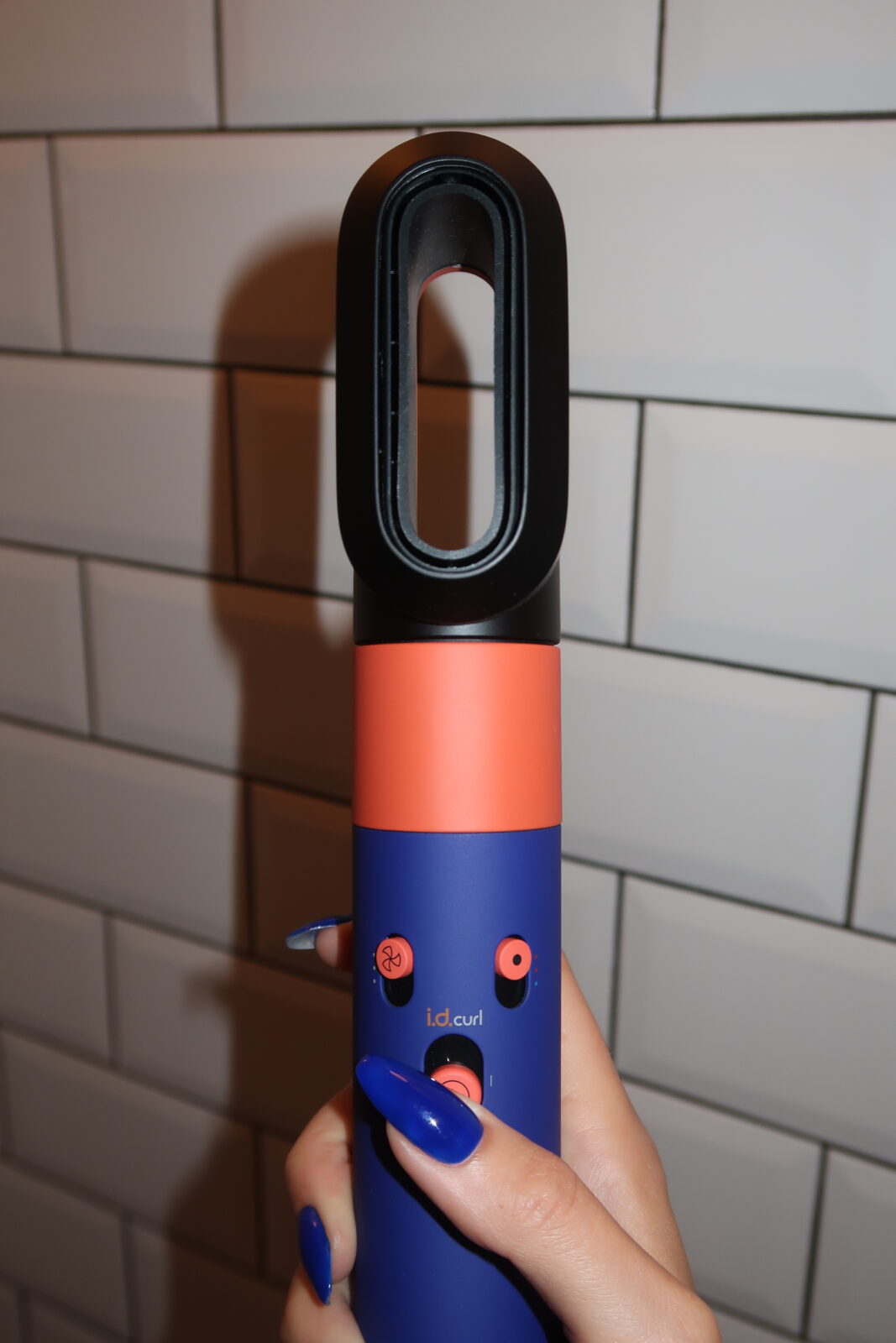 Multistyler Dyson Airwrap i.d.™ Straight+Wavy: Dyson Airwrap i.d.™ Straight+Wavy trzymany w dłoni na tle białych płytek. Widoczne przyciski sterowania, funkcja i.d. curl™ oraz charakterystyczne chabrowo-pomarańczowe wykończenie. Nowoczesny design i ergonomiczny uchwyt.