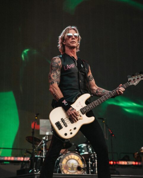 Basista z tatuażami i w kamizelce „L.A.M.F.” gra na białej gitarze basowej na scenie, z perkusją i logiem zespołu w tle.