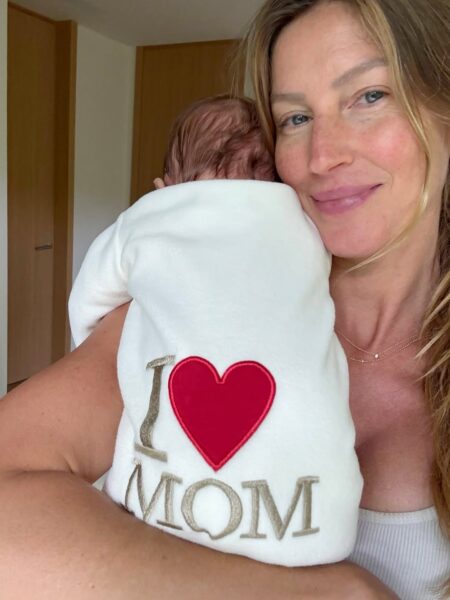 Uśmiechnięta kobieta trzyma na ramieniu niemowlę ubrane w biały kocyk z czerwonym sercem i napisem 'I ❤️ MOM'. Twarzy dziecka nie widać, a kobieta patrzy w obiektyw z czułością.