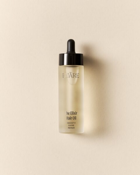 Szklana buteleczka z pipetą zawierająca olejek do włosów LUÃRE The Elixir Hair Oil. Minimalistyczne opakowanie z delikatnym, matowym wykończeniem, ustawione na beżowym tle. Produkt premium bez silikonów.