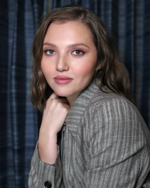 Zdjęcie kobiety w długich, ciemnoblond włosach z side part hair. Przedziałki mocno na bok to modne fryzury na jesień 2025. Kobieta pozuje półbokiem, oparta o blat, z ręką pod brodą. Ubrana jest w szary żakiet w paski.