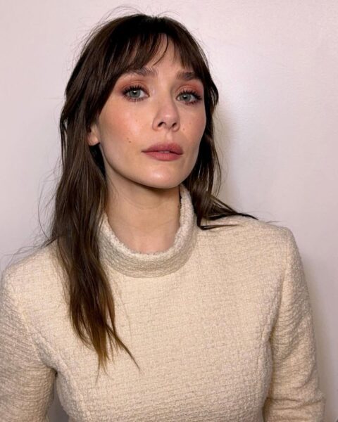 Zdjęcie aktorki Elisabeth Olsen w ciemnych, długich włosach z grzywką. Ubrana jest w beżowy sweter ze stójką. 