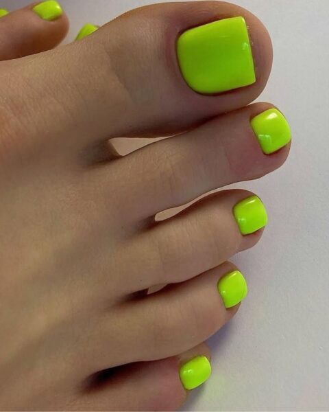 Zdjęcie stopy z efektownym pedicure z zadbanymi paznokciami pomalowanymi na kolor neonowej limonki. Stopa pozuje na białym tle.