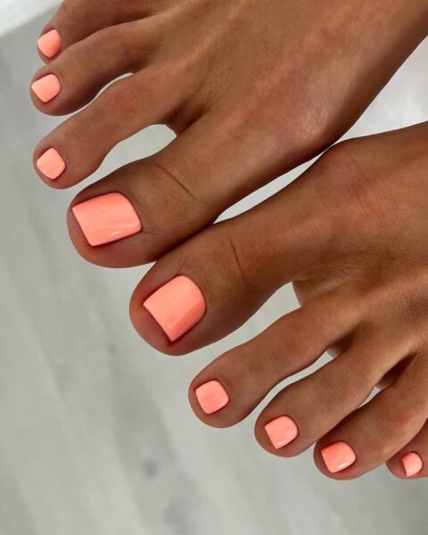 Zdjęcie stóp z efektownym pedicure w kolorze neonowej brzoskwini. Stopy są zadbane, tuż po pedicure i pozują tak, by prezentować stylizację.