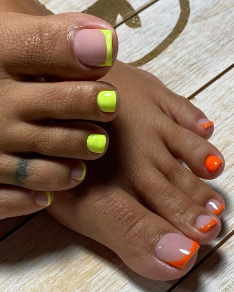 Zdjęcie stóp ze zbliżeniem na pedicure w neonowych kolorach: limonce i pomarańczu. Stopy pozują na tle drewnianej podłogi.