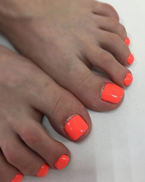 Zdjęcie stopy ze zbliżeniem na pedicure: zadbane, kwadratowe paznokcie pomalowane na neonowy pomarańczowy kolor. U nasady mają ozdobne kryształki. Stopa pozuje na białym tle.