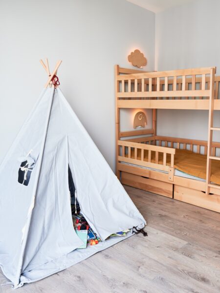 Jak wybrać łóżko piętrowe: Pokój dziecięcy z drewnianym łóżkiem piętrowym, wigwamem i eleganckim minimalistycznym wystrojem wnętrza w mieszkaniu po remoncie. Pokój dziecięcy z wygodnym łóżkiem dziecięcym, podłogą laminowaną, białymi ścianami i otwartymi drzwiami.