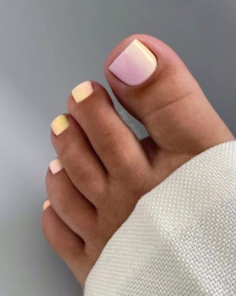 Zdjęcie stóp ze zbliżeniem na paznokcie u stóp, które mają zrobiony gradientowy pedicure w kolorze żółto-różowym.