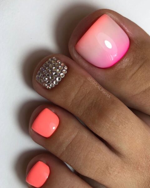 Zdjęcie stóp ze zbliżeniem na paznokcie z sunset ombre pedicure na lato 2025. Jest w kolorze pomarańczowo-biało-różowym, przez co wygląda jak zachód słońca. Jeden z palców jest ozdobiony srebrnymi cyrkoniami.
