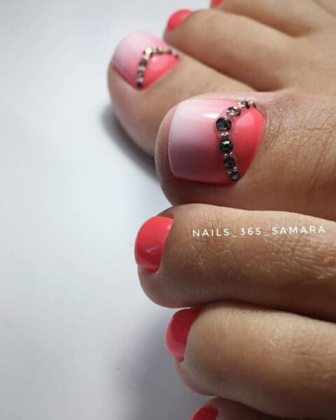 Zdjęcie stóp ze zbliżeniem na biało-czerwony pedicure w stylu ombre i dodatkowo ozdobiony małymi kryształkami. Stopy pozują na białym blacie.