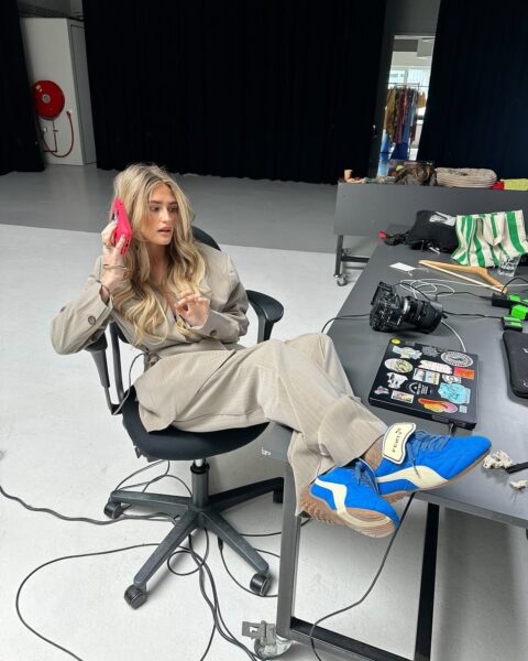 Kobieta w beżowym garniturze siedzi niedbale na krześle biurowym z nogami opartymi o stół, rozmawia przez różowy telefon; ma długie blond fale i nosi niebieskie sneakersy Puma x Fenty. Na stole przed nią leżą laptop z kolorowymi naklejkami, aparat fotograficzny i inne przedmioty studyjne.