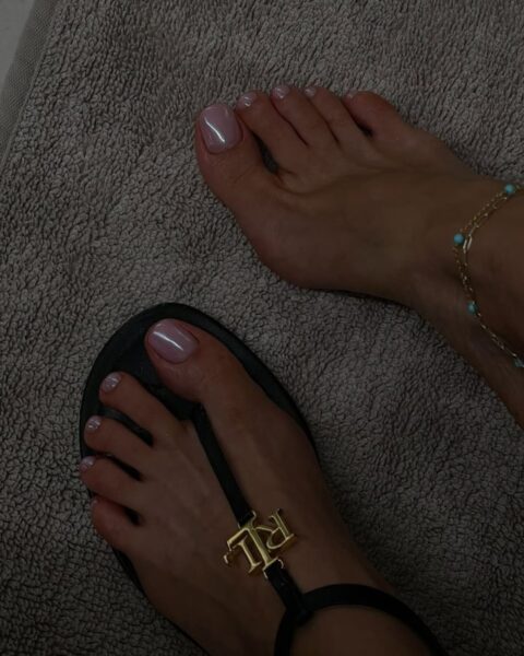 Zdjęcie stóp pozujących na tle szarego ręcznika. Widać opalowy pedicure w kolorze nude. Na jednej stopy jest sandał ze złotym loro marki, a na drugiej stopie bez buta jest bransoletka na kostce.