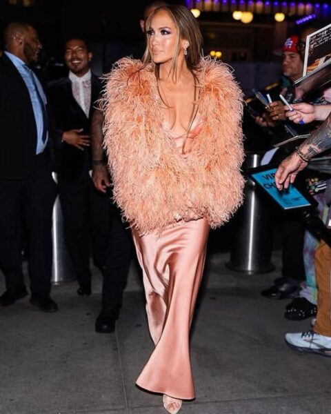 Zdjęcie Jennifer Lopez stojącej na ulicy wieczorową porą, a za nią widać ochroniarzy w garniturach. Sama jest ubrana w łososiową sukienkę maxi z satyny oraz brzoskwiniowe futro, a na nogach ma sandałki. Włosy proste, rozpuszczone oraz mocny makijaż.