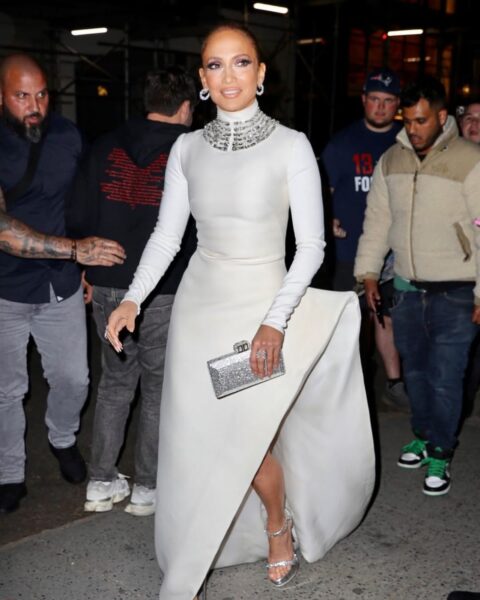 Zdjęcie Jennifer Lopez ubranej w długą, wieczorową suknię w kolorze srebrnej bieli ze stójką, długimi rękawami i charakterystycznym rozcięciem na nogę. Ma wysokie, srebrne sandały na platformie i prezentuje swój pedicure. Ma bogato zdobioną, wysadzaną kamieniami kolię oraz lśniące kolczyki w uszach. W ręce niesie srebrną torebkę typu puzderko. Idzie ulicą wieczorową porą, a za nią widać ochroniarzy w paparazzi.
