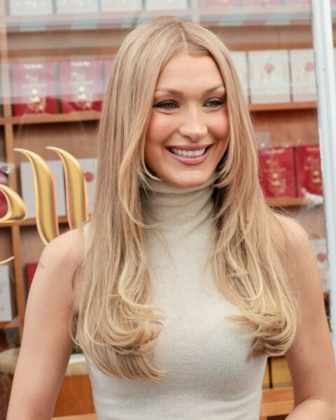 Zdjęcie modelki Belli Hadid w luksusowym butiki. Ubrana jest w beżowy top ze stójką i bez rękawów. Ma długie, rozpuszczone na ramiona blond włosy. Uśmiecha się szeroko.