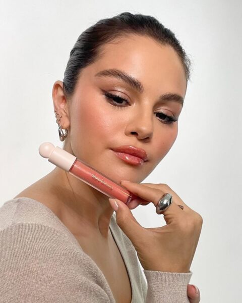 Zdjęcie Seleny Gomez, która pozuje z błyszczykiem swojej marki Rare Beauty. Ma ciemne włosy, gładko związane oraz rozświetlający makijaż. Ubrana jest w beżowy sweter. Pozuje półbokiem, ale patrzy w obiektyw. W ręce, przy twarzy, trzyma błyszczyk.