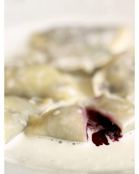 Zbliżenie na pierogi z jagodami polane śmietaną, jeden z pierogów lekko pęknięty, ukazując ciemnofioletowy, soczysty farsz.