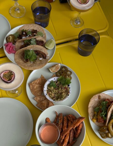 Stół z intensywnie żółtym blatem zastawiony kolorowymi daniami kuchni meksykańskiej. Na talerzach widoczne są tacos z mięsem i warzywami, nachosy z guacamole, frytki z batatów z sosem, a także koktajle z owocami passiflory. Obok stoją szklanki z wodą. Scena przedstawia letni, towarzyski posiłek w luźnej, kolorowej atmosferze.