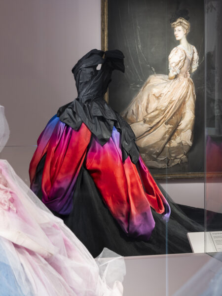 Dior wystawa 2025: Zachwycająca suknia haute couture o dramatycznym fasonie i kolorystyce przechodzącej od purpury przez intensywną czerwień po czerń, prezentowana w gablocie wystawowej. Projekt nawiązuje do kształtów kielichów kwiatowych, z charakterystycznym drapowaniem materiału w górnej partii. W tle wisi portret kobiety w balowej sukni z epoki, co podkreśla dialog między klasycznym pięknem a współczesną reinterpretacją.