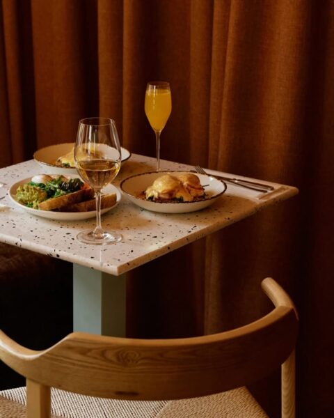 Elegancko nakryty stolik w restauracji z trzema talerzami śniadaniowymi — m.in. jajka w koszulce na toście z awokado i jajka po benedyktyńsku — oraz dwoma kieliszkami z napojami: białym winem i mimosa. W tle grube, brązowe zasłony, a z przodu widoczna drewniana krzesło z plecionym siedziskiem.
