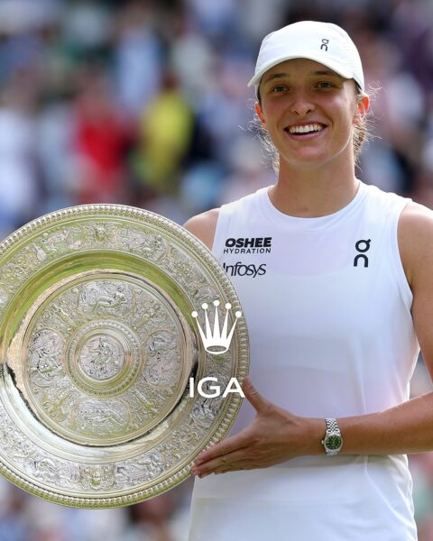 Iga Świątek stoi z triumfalnym uśmiechem na Centre Court w teledyskowej bieli i czapce, trzymając srebrną paterę — Venus Rosewater Dish — po swoim pierwszym zwycięstwie na Wimbledonie, które przypieczętowało jej szósty tytuł Wielkiego Szlema. W tle rozmyty tłum widzów.