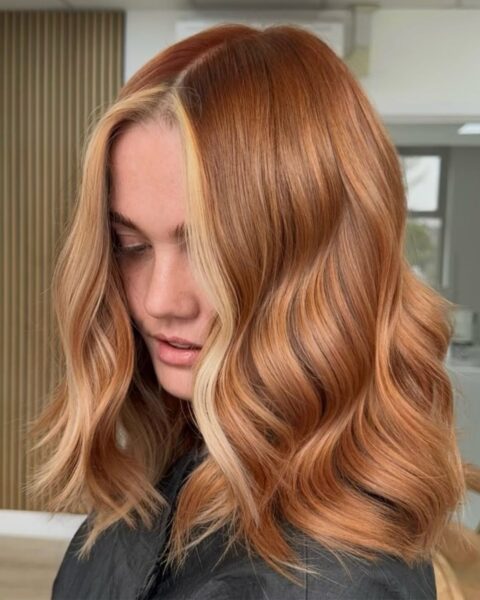 Zdjęcie kobiety w długich, rudych włosach, która ma refleksy strawberry blonde highlights. Ubrana jest w czarny fartuch fryzjerski. Pozuje w salonie.